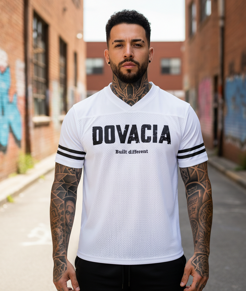 Man wearing a white 'DOVACIA' jersey with tattoos on arms in an urban setting